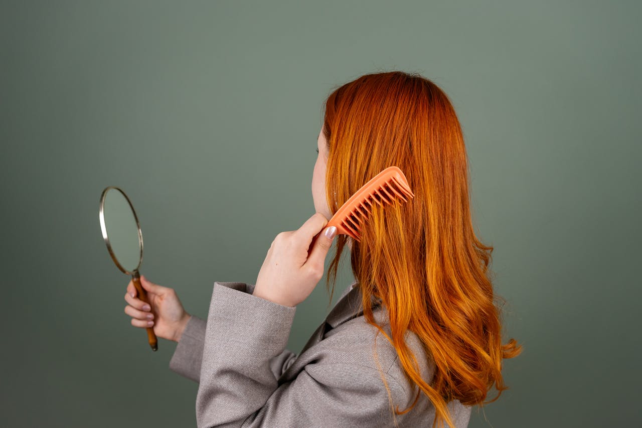 our-experience Red-haired woman using a comb while holding a mirror, focusing on hair care and beauty.